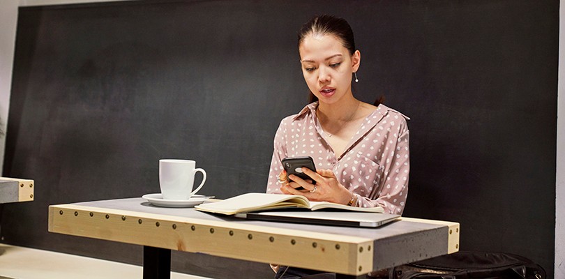 A woman sitting at a small cafe table with her laptop and enjoying a coffee, while looking down to the phone in her hands.