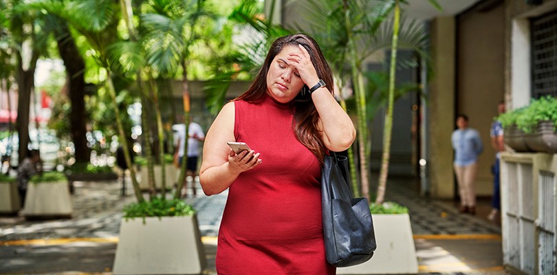 A woman in a red dress walking outside, brushing her hair back with one hand while she looks down at her phone in the other. 