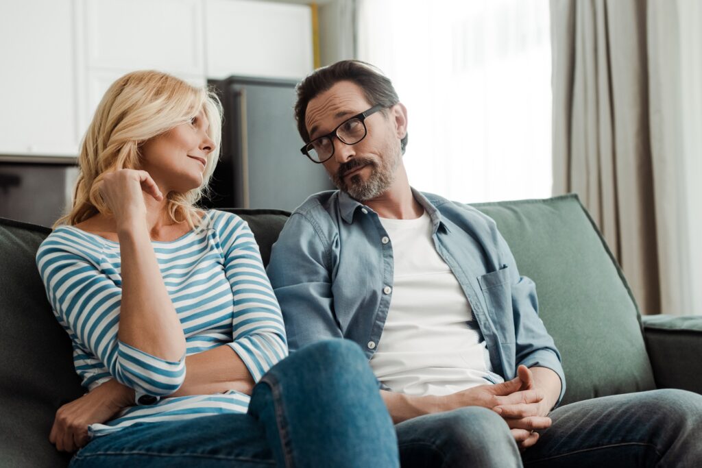 Mature man and woman sitting on a couch looking at each other with doubtful experessions